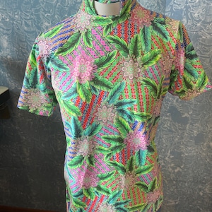 May include: A short-sleeved shirt with a vibrant floral pattern. The shirt features a high collar and a colorful design with pink, green, red, and blue elements. The fabric is covered in a repeating pattern of flowers and leaves.