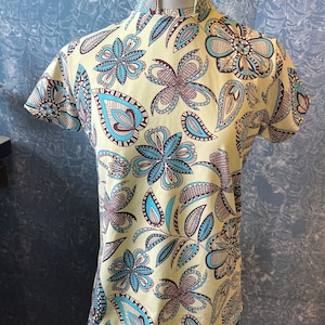 May include: A short-sleeved shirt with a high collar, featuring a cream-colored background and a repeating floral pattern in shades of teal, brown, and blue. The shirt is displayed on a mannequin.