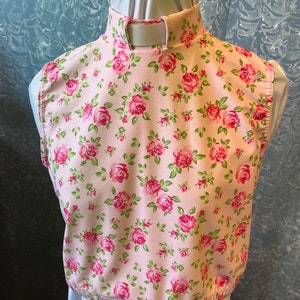 May include: A pink floral print bib with a high collar, featuring a pattern of red roses and green leaves. The bib is displayed on a white form, showcasing the design and shape of the garment.