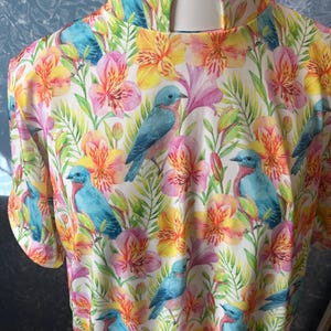 May include: A short-sleeved shirt with a vibrant floral and bird pattern. The design features blue birds, pink, yellow, and orange flowers, and green leaves on a white background. The shirt has a high, rounded collar.