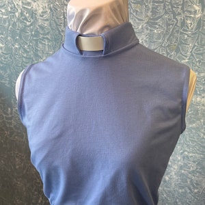 May include: A light blue clergy collar shirt with a white collar insert. The shirt has a high, rounded collar and a sleeveless design. The fabric appears to be a soft, breathable material.