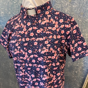May include: Navy blue short-sleeved shirt with a high collar, adorned with a pink floral pattern. The shirt features a white collar insert and a delicate floral design, creating a visually appealing garment.
