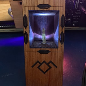 May include: A wooden box with a glass front, showcasing a small white figurine inside. The box is lit from within, casting a soft glow on the figurine. The box has a black triangle symbol on the front.