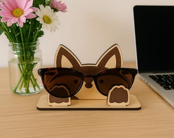 May include: A wooden eyeglasses holder in the shape of a fox with brown sunglasses resting on it. The holder is on a wooden desk, with a laptop and a vase of flowers in the background.