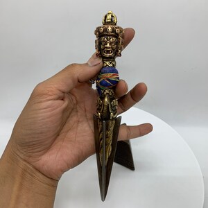 May include: A gold and copper colored metal dagger with a blue and red fabric wrapped around the handle. The dagger has a decorative top with a face and a sharp point.