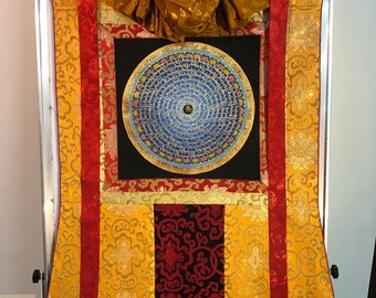 Puede incluir: Una rueda de oración tibetana enmarcada con letras doradas sobre fondo azul. El marco está decorado con tela roja y dorada.