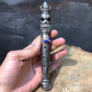 May include: A silver-colored, intricately carved, decorative object with a skull design at the top. The object has a blue and gold band around the middle and is held in a hand.