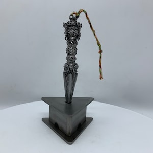 May include: A silver metal ritual dagger with intricate carvings and a decorative top. The dagger is on a triangular stand and has a colourful string attached to the top.