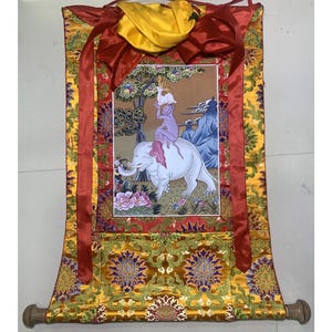 以下が含まれることがあります： 金色と赤色の縁取りが施された、伝統的なアジアの絵画が描かれたカラフルな絹の巻物。白い象に乗った人物が描かれています。巻物の上下には木製の棒が付いています。