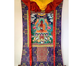 May include: A colorful Tibetan Thangka painting depicting a seated Buddha figure with a golden halo. The painting is framed with a red and gold border and is mounted on a blue and gold fabric scroll.