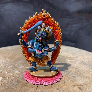 Puede incluir: Figura detallada de una deidad con tez azul, sosteniendo una espada y un cuenco. La figura está rodeada por un halo de fuego naranja y rojo. Se encuentra sobre una base rosa y dorada. La estatua mide unos 15 cm de altura.