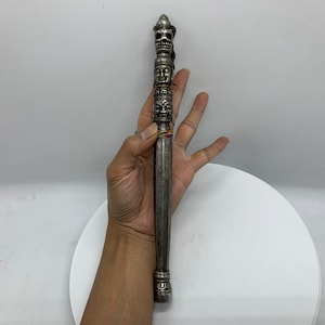 Puede incluir: Una varita de metal de color plateado con intrincados tallados de caras y calaveras. La varita se sostiene en una mano sobre un fondo blanco.