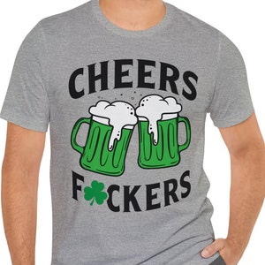 Op de afbeelding: Een heidekleurig T-shirt met een zwarte afbeelding van twee groene bierglazen met de tekst "CHEERS F*CKERS" en een groene klaver.