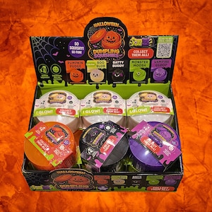 May include: A retail display box of Halloween Dumpling Squishies featuring six round containers in orange, black, and purple. The packaging includes graphics of a pumpkin, ghost, bat, monster, and vampire, with text reading Collect Them All.