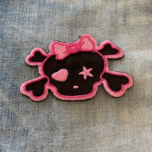 May include: Pink and black embroidered skull patch with a pink bow on top. The skull has a single star in one eye socket.