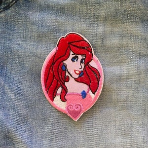 Può includere: Toppa ricamata raffigurante un personaggio dei cartoni animati con capelli rossi, orecchini blu e un vestito rosa. La toppa è bordata di rosso e posta su una forma ovale rosa chiaro. La toppa è su uno sfondo di denim blu.