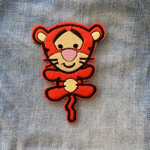 May include: Orange and black embroidered iron-on patch of a cartoon tiger with a smiling face. The tiger is sitting with its paws crossed and has a long tail.
