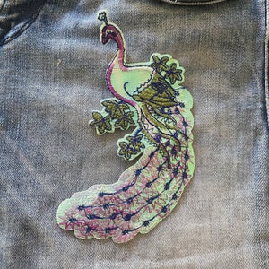 May include: A green and purple embroidered peacock patch with a detailed design. The peacock is standing on a branch with leaves and flowers.