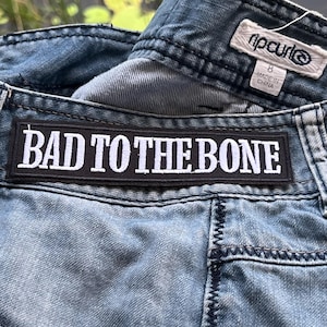 Peut inclure: Un jean en denim bleu avec un patch noir sur lequel est inscrit "BAD TO THE BONE" en lettres blanches. Le jean porte une étiquette de marque "Rip Curl" et "Made in China".