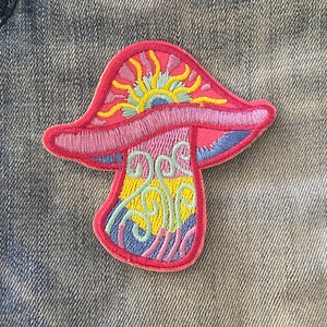 Pode incluir: Remendo de cogumelo bordado com uma tampa rosa, brânquias azuis e um desenho de sol amarelo e laranja. A haste apresenta redemoinhos amarelos, azuis e roxos. O remendo está em um fundo de ganga.