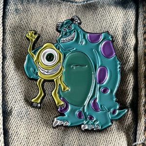 Op de afbeelding: Een blauw en paars monster met één groot oog en een geel monster met drie ogen. De monsters lachen en lijken vrienden te zijn. De afbeelding is een pin.