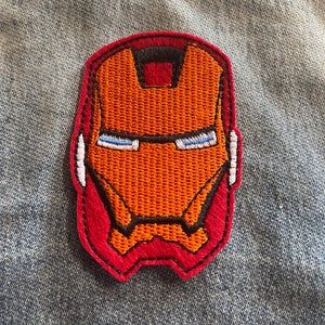 Puede incluir: Parche bordado con el diseño de la máscara de Iron Man. El parche tiene una cara naranja con detalles en negro y blanco, delineada en rojo. El parche está cosido en una chaqueta vaquera.