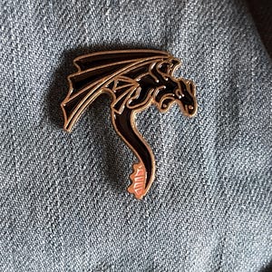 Könnte beinhalten: Schwarzer Emaille-Drachen-Pin mit goldener Umrandung und rosa Schwanz. Der Pin befindet sich auf einem blauen Jeans-Hintergrund.