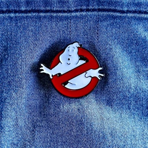 May include: A red and white enamel pin featuring the Ghostbusters logo. The logo is a red circle with a diagonal line through it, and a white ghost with a surprised expression is inside the circle.