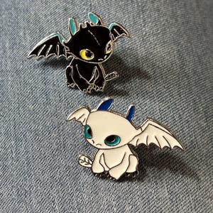 Może przedstawiać: Dwie emaliowane przypinki z motywem smoków. Jedna przypinka jest czarna z żółtymi oczami i turkusowymi akcentami, druga biała z turkusowymi oczami i niebieskimi akcentami. Obie przypinki mają srebrne skrzydła i obramowania.