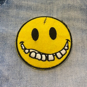May include: Yellow smiley face patch with a stitched-up mouth and crooked teeth, sewn onto a blue denim fabric.