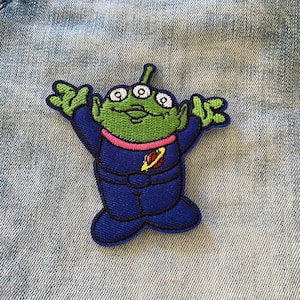 May include: Green alien character with three eyes, wearing a blue spacesuit with a pink collar and a red planet design. The alien is smiling and has its arms outstretched.
