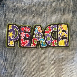 May include: A colourful embroidered patch with the word "PEACE" in a variety of fonts and designs. The patch is sewn onto a blue denim fabric.