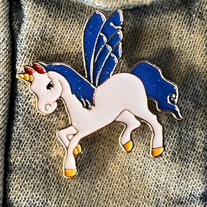 May include: A gold-colored enamel pin featuring a white unicorn with blue wings and a red horn. The unicorn is standing on its hind legs with its front legs raised.
