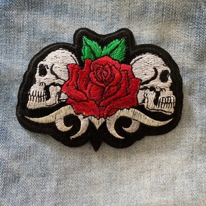 Może przedstawiać: Haftowana naszywka z czerwoną różą i dwiema białymi czaszkami. Naszywka ma czarną obwódkę z dekoracyjnymi wirami i zielonymi liśćmi. Do odzieży lub akcesoriów.