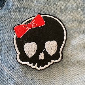 May include: Black skull patch with white eyes and a red bow. The skull is outlined in white and has a heart shape for each eye.