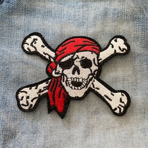 Op de afbeelding: Een witte schedel en gekruiste beenderen patch met een rode bandana. De patch is geborduurd met zwart garen en heeft een witte achtergrond.