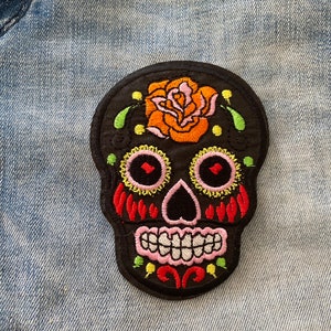 May include: A black skull patch with colorful embroidery. The design features a rose on top, pink and yellow accents around the eyes, and red details on the cheeks. The patch is placed on a light blue denim fabric.