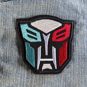 May include: A black, white, red and teal embroidered patch of the Transformers logo. The logo is a stylised robot face with a black background.