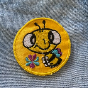 May include: A yellow embroidered patch with a cartoon bee holding a flower. The bee has black eyes, a black and yellow striped body, and a black antenna. The flower is purple and blue.