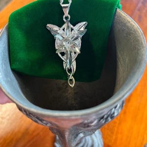 May include: A silver-toned pendant necklace featuring a star-shaped design with clear crystal accents. The pendant is suspended from a delicate chain and rests on a green velvet surface. The necklace is displayed inside a decorative metal chalice.