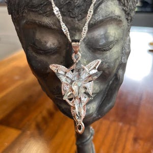 May include: A silver pendant necklace with a star-shaped design and clear crystal detailing. The pendant hangs from a delicate chain. The necklace is displayed on a decorative chalice.