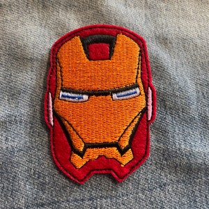 Peut inclure: Visage d'Iron Man brodé sur un patch rouge avec des détails orange et noirs. Le patch est sur un fond en jean bleu.