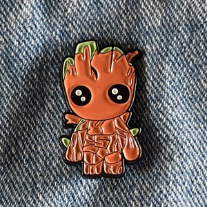 以下が含まれることがあります： ガーディアンズ・オブ・ギャラクシー映画シリーズのキャラクター、グルートの漫画風キャラクターが描かれた、茶色と緑のエナメルピン。キャラクターは腕を体の横につけた状態で立っており、大きく丸い頭と大きな目が2つあります。