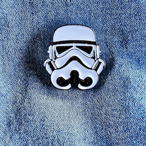 May include: A white enamel pin of a Star Wars Stormtrooper helmet. The pin is on a blue denim background.