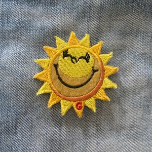 Puede incluir: Un sol amarillo bordado con una cara sonriente y un ojo guiñando. El sol tiene un contorno rojo y naranja y está sobre un fondo de mezclilla azul.