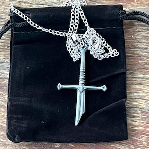 May include: A silver-toned metal pendant shaped like a broken sword, suspended from a matching silver-toned link chain. The pendant features a detailed hilt and a jagged, fractured blade edge, resting against a black fabric drawstring pouch.
