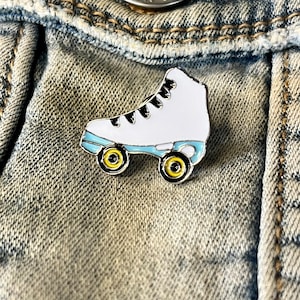 Könnte beinhalten: Weißer Rollschuh-Emaille-Pin mit schwarzen Schnürsenkeln und gelben Rädern. Der Pin befindet sich auf einem blauen Jeans-Hintergrund.