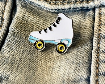 Könnte beinhalten: Weißer Rollschuh-Emaille-Pin mit schwarzen Schnürsenkeln und gelben Rädern. Der Pin befindet sich auf einem blauen Jeans-Hintergrund.