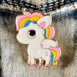 May include: A white enamel pin with a cartoon unicorn design. The unicorn has a pink, yellow, and blue mane and tail. The unicorn has a black eye and a white body.