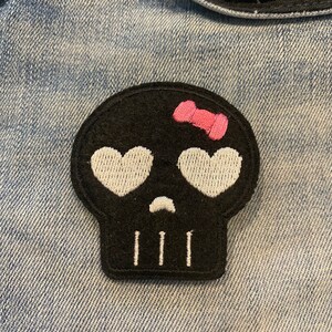 May include: Black skull patch with white eyes and a pink bow. The skull has a small frown and three white teeth.